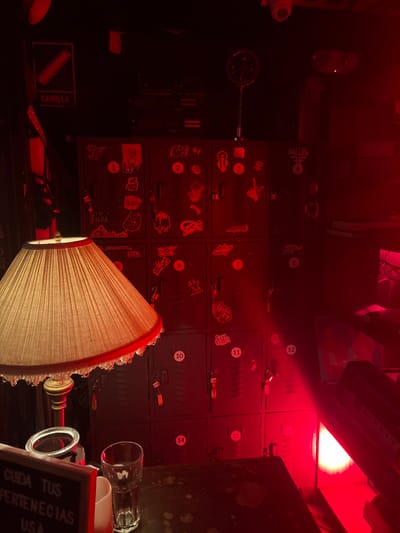 The scene depicts an interior space within a commercial establishment, likely a nightclub or bar, located in Bogota, Capital District, Colombia. The environment is intensely illuminated by pervasive red lighting, casting all objects in a deep crimson hue and creating strong contrasts.

In the center-right, a bank of fifteen dark-colored metal lockers is arranged in three columns and five rows, numbered sequentially 1 through 15 with white circular labels. Numerous small padlocks with keys attached hang from the handles of most lockers. The locker surfaces are heavily adorned with a variety of stickers, including stylized text, skulls, and abstract designs.

In the foreground, to the left, a table lamp with a pleated, light-colored fabric lampshade featuring a red fringe and clear dangling ornaments is positioned on a dark, possibly stained, tabletop. Adjacent to the lamp, a partially visible white sign board displays Spanish text: "CUIDA TUS PERTENECIAS" (Care for your belongings) and "USA." A clear glass and a white cylindrical object are also present on the table.

Above the lockers, mounted on a dark wall, a white sign with a red border and a lever icon indicates "CAMILLA" (Stretcher). Higher up, an overhead security camera (CCTV) is visible, along with other mounted equipment, including a circular object on a flexible arm. A concentrated source of bright red light emanates from the lower right portion of the scene. No individuals are present. The setup suggests a designated area for patrons to store personal items.