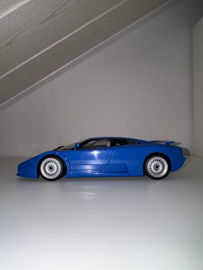 A blue toy car is wedged vertically in a corner. The wheels are silver with black tires. The car's body is shiny, reflecting light. The background consists of two white walls meeting at a right angle. A section of a wooden structure with slats is visible on the left, providing a contrasting texture to the smooth walls.