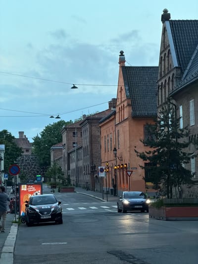 The image depicts a street scene in Oslo, Norway, likely in a historic district, with traditional brick buildings lining both sides of the road. The sky is overcast, suggesting it is either early morning or late evening.

On the left side of the street, a dark-colored electric car, identified by the license plate "EV 73135" and "GSS" branding, is parked parallel to the curb. Next to it is a large advertising billboard with text in Norwegian, which translates to "We are back again!" featuring an image of a soccer ball and cookies. A person is walking away from the billboard towards the left edge of the frame. A blue and red circular parking sign with a diagonal red line is also visible on the left.

The street itself is paved and appears to be a two-way road with marked pedestrian crossings. On the right side of the street, another dark-colored electric car, with the license plate "EV 62662", is driving towards the viewer with its headlights on. The building on the right is a multi-story brick structure with arched windows. There is an orange-colored building on the right side of the street further down, featuring a steeply pitched dark tiled roof and decorative gables topped with finials.

Street lamps are mounted on poles along the sidewalk and overhead wires with hanging lights are strung across the street. Trees are present along the street, with a dense evergreen tree visible on the right side, partially obscuring one of the buildings. In the background, older stone structures and a tower are visible, hinting at the historical significance of the location. The overall impression is of a well-preserved urban environment with a blend of historical architecture and modern elements like electric vehicles and digital advertising.
