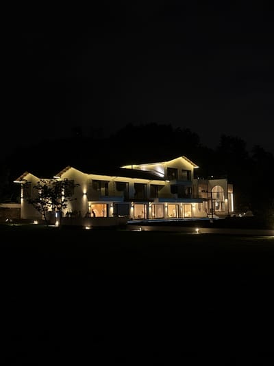 A multi-story white building is illuminated at night, with lights on both the exterior and visible within the windows. The building is situated between dark trees and a dark grassy area. The location is likely in Jinhua, China, given the request. No people are directly visible, though there are indications of occupancy through interior lights. The environment is dark, with artificial light sources providing illumination.