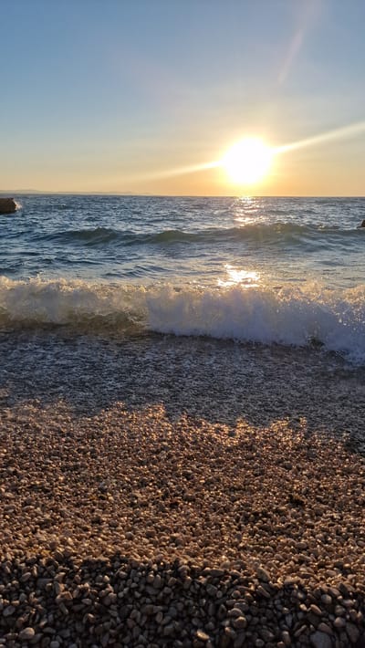 This is a vertical, wide-angle photograph of a pebble beach meeting the sea at sunset. The sun, a brilliant orb with intense rays, dominates the left side of the frame, casting a warm golden light across the scene. The sky transitions from a pale blue at the top to a soft yellow-orange near the horizon. Gentle waves are breaking on the shore, with the foam catching the sunlight and appearing a sparkling white. The beach itself is covered in smooth, small pebbles of various shades of grey, brown, and beige, which are illuminated by the low-angle sun, creating a textured and shimmering surface. The overall mood is serene and picturesque, typical of a peaceful sunset at a coastal location. There is no visible text in the image. The location is Zadar, Croatia.