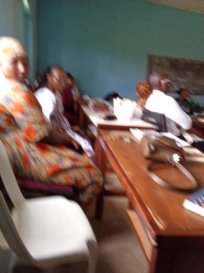 The image depicts an indoor scene, likely a classroom or meeting room, with several individuals present. The lighting suggests it is daytime. A woman in the foreground is seated, wearing a colorful dress with orange and blue patterns. To her right, another person, wearing glasses and a white top, is also seated. Further back, a person in a white garment is visible. The room has light blue walls and a wooden table in the foreground. On the table, there are papers, a purse or bag, and a book with blue lettering. The overall image is blurry, making it difficult to discern specific details or expressions. The setting appears to be in Kakuri Gwari, Nigeria, as indicated by the provided location context. There is no discernible activity or event taking place beyond people being seated at tables.