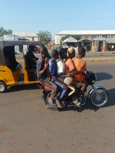 In Tumfure, Nigeria, a yellow auto-rickshaw is parked on the right side of the road. A man wearing a dark shirt is seated in the driver's position of the auto-rickshaw, and a person wearing a dark-colored shirt is also visible in the passenger seat. Five young men are riding on the back of a motorcycle. The driver of the motorcycle is wearing a blue shirt, and the other four passengers are wearing a white shirt, an orange shirt, and a light-colored shirt. One of the passengers is holding a white plastic container. The motorcycle is traveling on a paved road in front of buildings. The sky is clear and blue.