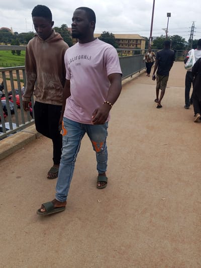 Two young men walk on a pedestrian overpass in Immigration Quarters, Nigeria. The man in the foreground, wearing a pink "California 1991" t-shirt and distressed blue jeans, walks towards the viewer, his right foot forward in a dark green slide sandal. He has a beaded bracelet on his left wrist. Beside him, the man on the left wears a tan hoodie and black pants, also walking forward, his left foot in a dark green slide sandal. They are on a dusty brown concrete walkway with a metal railing on their left, overlooking a street with parked cars below. In the background, other pedestrians are visible on the overpass, as well as buildings and trees. The sky is overcast.