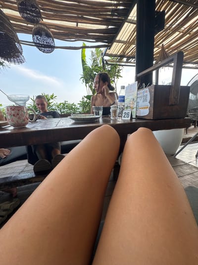 The image captures an outdoor dining scene from a first-person perspective, with the photographer's bare legs extending into the immediate foreground, resting on or near a rustic wooden table.
On the table, various items are visible: two clear plastic water bottles, multiple drinking glasses, a small white mug adorned with a red floral pattern, and a salt and pepper shaker. Information cards are placed on the table, one displaying "google map" with a QR code and a phone number beginning with "+84", and another with the word "WARNING". A small stand clearly shows the table number "26".
Two other individuals are seated at the table. To the left, a woman with dark hair tied back leans forward, appearing engaged with something on the table. Further down, another person is partially visible, also seated.
The setting is an open-air establishment, likely a cafe or restaurant in Hà Giang 1 Ward, Vietnam. It features a slatted bamboo or wooden roof structure providing shade, from which woven basket decorations are suspended. The background shows a clear, bright sky and lush green foliage, indicative of a warm, tropical climate. A ceiling fan is partially visible overhead. The bright daylight suggests the time is likely daytime, either morning or afternoon. The skin on the visible legs appears fair with a few small red marks.