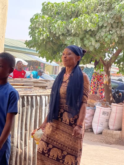 An outdoor market scene is depicted in Samaru, Nigeria, during daylight hours. In the mid-ground, an adult female with dark skin is positioned slightly to the right of the frame's center, looking upward and to the right with her mouth slightly open. She wears a black headwrap, a black sheer scarf, and a long brown and gold dress patterned with elephant motifs. A pink beaded necklace is visible, along with an orange/red beaded bracelet on her left wrist. She holds a yellow and white plastic item in her left hand at hip level.
To her immediate left, in the foreground, the left side of a young male's head and blue t-shirt are partially visible. Behind the main female subject, an adult male in a red t-shirt with a white logo is visible in the mid-background, looking right. Further left and behind him, another adult male in a bright blue t-shirt and a dark cap leans over a corrugated metal and wooden structure, possibly a market stall.
To the right of the main female subject, a large tree with green foliage stands. Hanging from a line near the tree are various patterned clothing items. At the base of the tree, multiple large white sacks, two marked "DEFTA 100", contain what appears to be agricultural produce. A motorcycle is partially visible behind these sacks. The background features several market stalls with corrugated roofs, other individuals, and parked vehicles, indicating a lively commercial area. The sky above is clear, suggesting bright weather conditions.
