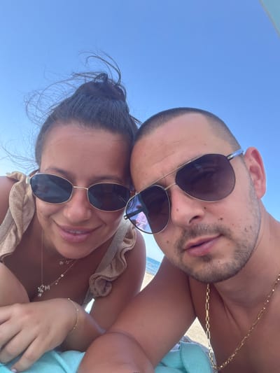 A close-up, low-angle selfie captures a young couple lounging on a beach under a clear blue sky. The woman, on the left, has dark hair pulled up in a bun and is wearing a gold sparkly top with ruffled straps and gold sunglasses. She is smiling and looking at the camera. A delicate gold necklace with a small pendant and a bracelet adorn her neck and wrist. The man, on the right, has a buzzed haircut and is wearing aviator-style sunglasses. He has stubble and is wearing a gold chain necklace. The background shows the ocean and beach. The overall impression is one of a casual, romantic day at the beach.