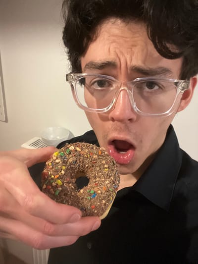 A man with dark hair and clear glasses holds a chocolate donut covered in sprinkles. His mouth is open, tongue slightly visible, appearing ready to eat the donut. He wears a black shirt. The background shows a white wall and a partial view of a white object. The picture is rotated.