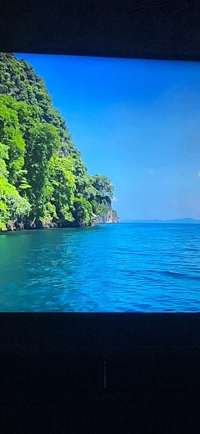 The image depicts a lush, green island or cliff face jutting into a vibrant blue ocean under a clear blue sky. The vegetation on the landmass is dense and appears to be tropical. The water is calm and reflects the sky. There are no people or man-made objects clearly visible in the scene, apart from what might be some small structures nestled into the foliage on the side of the cliff. The scene is a natural landscape. There is no information in the image to suggest a location in Bila Tserkva, Ukraine.