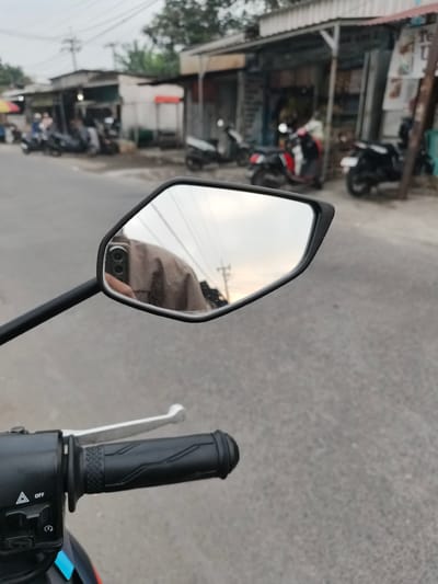 The image is taken from the perspective of someone on a motorcycle, looking into the left-side mirror. The mirror reflects the rider's torso, wearing a light brown shirt, and their hand holding a mobile phone, which has a dual-camera setup. The background visible in the mirror shows a hazy sky with some clouds, suggesting it might be late afternoon or early morning.
The foreground of the image shows the motorcycle's handlebar, with a black grip and a silver brake lever. A control panel with buttons and labels like "OFF" and a hazard light indicator is also visible.
Outside the mirror's reflection, the environment appears to be a street in a city or town. On the right side of the road, there are several small shopfronts with corrugated metal roofs. Parked motorcycles are visible in front of these shops. The road itself is paved with asphalt.
Based on the context provided that the location is Sidorejo, Indonesia, this scene likely depicts a street in that Indonesian city, potentially near a marketplace or a row of local businesses. The presence of multiple motorcycles indicates a common mode of transportation in the area. The overall atmosphere is casual and urban.