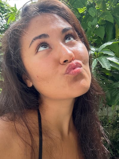 A close-up, sideways selfie of a young woman with long, dark hair is captured against a backdrop of lush green foliage, suggesting an outdoor setting in a tropical location like Peleana, Sri Lanka. The woman's face is partially visible, with her eyes looking to the left and her lips pursed in a playful pout. Her skin appears smooth and sun-kissed, with a few faint freckles visible on her cheek. Her dark eyebrows are neatly shaped, and her eyelashes are long and full, framing her expressive eyes. Her lips are full and slightly parted, with a subtle sheen that suggests lip balm or gloss. The lighting appears natural, likely from daylight, casting gentle shadows and highlighting the textures of her skin and hair. The leaves in the background are vibrant and detailed, indicating a bright, possibly sunny, day. The overall mood is casual and relaxed, with a hint of flirtatiousness conveyed through her pose and facial expression. There is no visible text in the image.