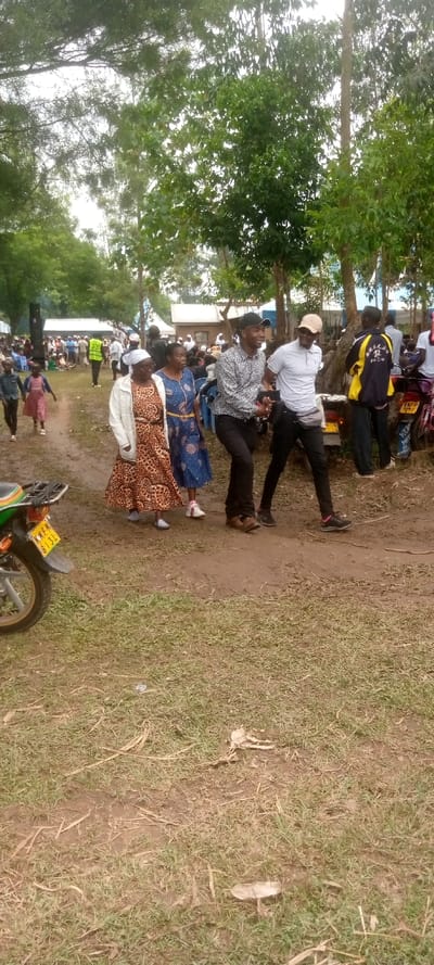 The image depicts a scene in Burundu, Kenya, a rural village situated at the coordinates latitude 0.459, longitude 34.776. The scene takes place on a dirt road lined with green grass. Four individuals are walking along the road, two women and two men. The women are dressed in traditional clothing with patterned dresses and head coverings. The men are wearing modern casual attire. They are walking toward a larger group of people in the background, likely attending a social event or gathering. The foreground includes a parked motorcycle, and the background features various buildings and trees, suggesting a typical Kenyan village setting. The atmosphere is serene and peaceful, reflecting a common rural scene in Kenya.
