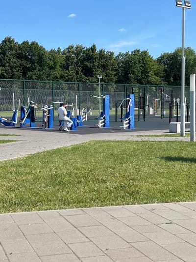 This media file depicts an outdoor gym scene at a park or recreational area, likely in Tolyatti, Russia. The foreground features a paved walkway bordering a well-maintained grassy lawn. Beyond the walkway, a chain-link fence encloses a series of blue and white outdoor fitness equipment. One person, dressed in a white t-shirt and jeans, is actively using a weight machine, positioned on the equipment and appearing engaged in their workout. Another person, wearing a grey shirt and shorts, is visible on a different piece of equipment, also seemingly exercising.
The setting is bathed in bright daylight, suggesting it's daytime. The sky is clear and blue with a wispy white cloud in the distance. Lush green trees form a dense backdrop behind the gym equipment and fence. The overall impression is one of a pleasant, active day in a public outdoor space designed for physical fitness. The equipment appears to be designed for a variety of exercises, with visible handles, weights, and seating. The scene is peaceful and functional, focused on exercise and recreation. No visible text is present in the image.