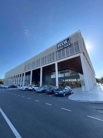 The image shows the HDL Retail Park located in Bjeliši, Montenegro, under a clear blue sky with a few wispy clouds. The modern building features a distinctive angled design with a white perforated facade and large white support columns. The HDL Retail Park logo is prominently displayed on the upper facade. In front of the building, there is a parking lot filled with various cars. The foreground shows a dark asphalt road with white lane markings. The environment appears to be a commercial area with some greenery visible in the background and to the right. The sun is visible in the upper right portion of the image, creating a lens flare effect. The scene is static and depicts a typical day at a retail park.