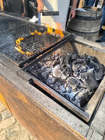 A close-up shot captures a grilling setup on what appears to be a sunny day, likely in Óbidos, Portugal. In the foreground, a deep metal container is filled with charcoal, some of which is glowing red beneath a layer of ash. Above the charcoal, a metal grate is positioned, with flames licking upwards in a heart-shaped pattern. A person, wearing a light-colored apron over blue jeans, stands to the right, holding a metal brush in their right hand, seemingly in the process of cleaning or tending to the grill. To the far right, a large wooden barrel, typical of those found in wine regions, is visible, along with the legs of several individuals, also dressed casually. The ground beneath the setup is paved with light-colored cobblestones. The overall impression is of an outdoor food preparation area, possibly at a market or restaurant in Óbidos.