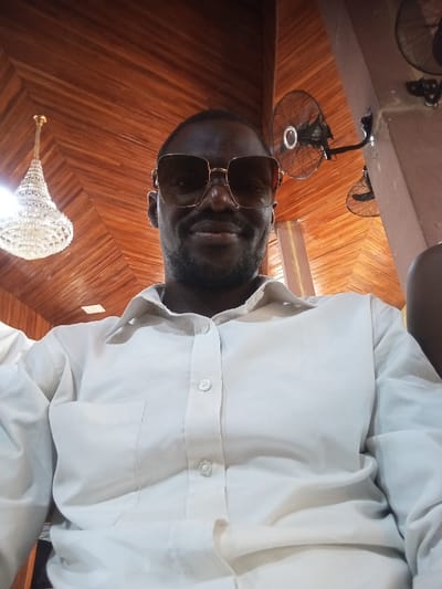 This is an eye-level indoor portrait captured in the city of Ogun-Aja, Nigeria. The primary subject is a dark-skinned adult male, positioned centrally, looking directly at the camera with a faint smile. He wears large, rectangular, gold-framed dark sunglasses and a light beige or off-white collared button-down shirt with a visible left chest pocket. His short hair and beard are dark. The background features a sloped or vaulted ceiling constructed from reddish-brown wooden panels. To the left, a large, ornate chandelier composed of numerous clear or frosted beads hangs, appearing illuminated. To the right, mounted on a dark vertical structural element, are two black wall-mounted oscillating fans; one is fully visible with a light-colored central hub, and the other is partially cropped.