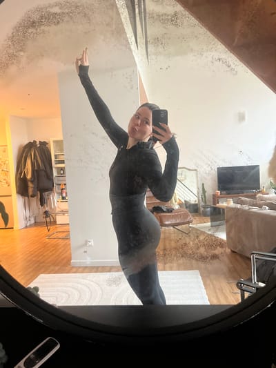 The image displays a female individual, dressed in a black, long-sleeved athletic top and matching leggings, captured in a selfie taken via an oval-framed mirror within a residential interior in Roubaix, France. The subject stands with her right arm extended upwards and her left arm holding a dark-colored smartphone, aimed towards her face, which exhibits pursed lips. The mirror itself shows considerable de-silvering and dark, irregular mottling, predominantly across its upper and lower sections and reflecting the floor, which causes localized visual distortion.

The reflected environment reveals a multi-room dwelling. To the subject's left, a light-toned wooden floor leads to a white wall with multiple dark jackets hanging on hooks. Further left, a warm light source illuminates an area containing a white open-shelving unit with various objects and a transparent plastic chair with a chrome frame. A power outlet with a white cable is visible on this wall.

To the subject's right, the reflection extends into a living area. A large, rectangular white rug with a circular pattern covers a section of the wooden floor. Furnishings include a dark brown leather armchair with a metal frame, a flat-screen television resting on a dark stand, and a light-colored sofa or sectional. A tall, slender plant in a dark pot is positioned behind the armchair. A staircase with dark railings is visible in the background, suggesting multiple levels. A smaller, decorative mirror is mounted on the wall behind the armchair, and a black folding chair is partially visible on the far right. The interior is brightly illuminated.