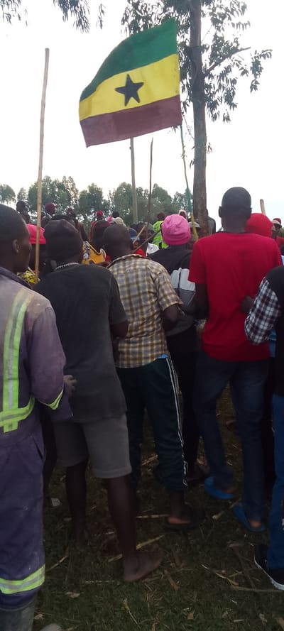 A group of men stand in a clearing in a grassy field. The men are mostly wearing casual clothes, including a few in plaid shirts and one in a red shirt. The men are standing in a group with their backs to the camera. Some of the men are wearing sandals, while others are barefoot. In the background, a Senegalese flag is being held up on a stick. There is a tall tree with leaves, which is slightly out of focus. The ground is covered in grass and dirt.  The location of the image is not in Kenya, as the provided coordinates correspond to a location in the Indian Ocean. It is not possible to determine the precise location based on the image.