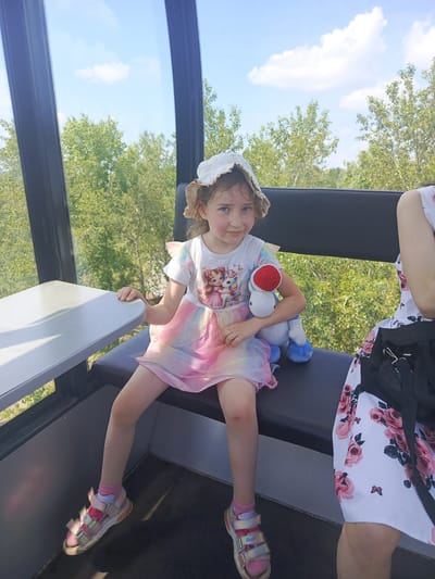 A young girl with blue eyes, curly brown hair, and a straw hat sits in a cable car. She is wearing a white t-shirt with a graphic of two girls and a unicorn, and a rainbow-colored skirt. She holds a blue and white stuffed toy with a red mushroom on its head. To her right, a portion of another person's arm and shoulder wearing a white dress with pink roses is visible. The cable car is moving, as indicated by the blurry trees and sky outside the windows. The interior of the cable car features a grey table and black seating. The location is the Angarsk Cable Car in Angarsk, Russia.