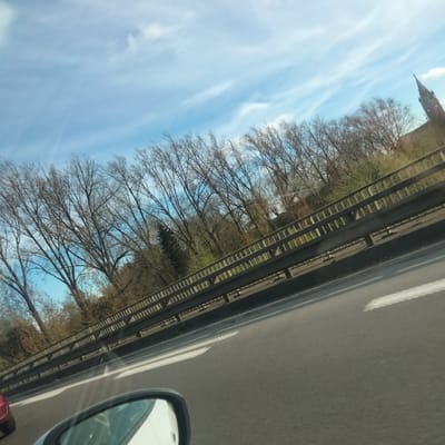 The image depicts a view from inside a vehicle, likely a car, traveling on a highway. A side mirror and part of the dashboard are visible in the foreground. The road stretches out in front, marked with white lane indicators. A metal guardrail runs alongside the road. Beyond the guardrail, there is a line of trees, mostly bare, with some hints of green foliage beginning to emerge, suggesting it's early spring. In the distance, on the right side of the image, a church steeple can be seen. The sky is blue with streaks of white clouds. Given the location provided, this is likely a road near Erquinghem-Lys, France.