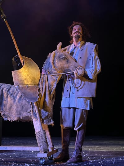 This image captures a theatrical stage scene in Novoshakhtinsk, Russia, featuring a male actor and a horse prop. The actor stands on the right, facing slightly left and upwards, with a focused gaze. He possesses long dark hair, a prominent dark mustache, and a pointed beard or goatee. His attire consists of a light beige long-sleeved shirt, layered with a matching sleeveless tunic featuring dark circular grommets along the front and shoulder seams. He wears dark trousers and dark lace-up boots. With both hands, he holds the fabric head of a stylized horse prop, which appears aged and distressed with a rope bridle.
The horse prop is constructed with a visible wooden frame at its base, wrapped with rope and equipped with small caster wheels. Its body is covered in rough, light brown fabric, possibly burlap, with torn edges. Mounted on the horse's back is a metallic, golden-hued piece of armor or a saddle component with a jagged, irregular bottom edge. A long, slender spear or lance, featuring a metallic tip or blade, stands upright behind the horse prop, partially visible on the left side of the frame. The background is completely black, indicating a darkened stage. The stage floor is dark, strewn with numerous small, light-colored pieces of debris. The scene is illuminated by theatrical lighting, casting warm light on the subjects and cooler light on the stage floor.