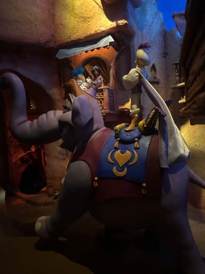 An indoor scene, likely from a theme park attraction, depicts animatronic figures set in a Middle Eastern or Arabian environment. The main subject is a large, light purple or grey elephant, decorated with a blue and gold ceremonial saddlecloth featuring heart-like motifs. On the elephant's head, a smaller, monkey-like figure, possibly Abu, is perched, holding a white object. Another human-like figure, resembling Aladdin, is seated on the elephant's back, wearing a white top and blue pants, and appears to be playing an instrument or holding an object near its mouth. A gold Genie's lamp is prominently displayed on the elephant's saddle.
The background consists of multi-level buildings with textured, sandy-colored walls, arched doorways, and latticed windows, suggesting a bustling marketplace or town square. Several other figures are visible within these structures; one figure resembling Jasmine is seen leaning out of a window in the mid-ground. The lighting is dim and theatrical, with warm light illuminating the foreground and cooler blue light in the upper background, creating depth and atmosphere. The scene evokes a moment from Disney's Aladdin, likely from the "Le Passage Enchanté d'Aladdin" walk-through attraction or a similar ride at Disneyland Paris, given the location context of Chessy, France. No visible text is present in the image.