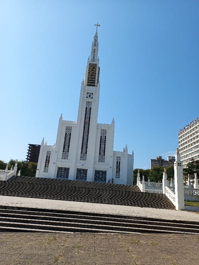 The image shows the Catedral de Maputo, a white, tall, rectangular church with a cross on top. It is located in the city of Maputo, Mozambique, at the coordinates -25.967, 32.573. There are a set of stairs in front of the church leading to a large paved area. The stairs and the paved area are empty. The sky is blue, and there are some trees visible on either side of the church. The church has large, rectangular windows that are made of glass. There are also a few other buildings visible in the background, including a tall, multi-story building with a flat roof on the right. The building is also mostly white, with black windows. The background is slightly blurred, but the church and its front steps are in focus.