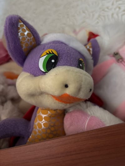 A plush toy is depicted in a close-up, angled view, positioned behind a reddish-brown wooden barrier that bisects the lower portion of the image. The toy is predominantly purple, with a cream-colored muzzle featuring two small black dots for nostrils and an orange fabric mouth. Its large, fixed eyes consist of black pupils, green irises, and yellow sclera. The pointed ears have inner panels with a silver fabric displaying orange circular patterns. A white, fuzzy band rests atop its head. Portions of its body are white and pink, with a red fabric detail visible behind its left shoulder. The immediate background is a blurred white, fluffy textile, while further to the right, a pink and white striped fabric and a silver zipper pull are discernible. Other indistinct, light-colored soft objects are present in the left background. The scene is located in Velikiye Luki, Russia.