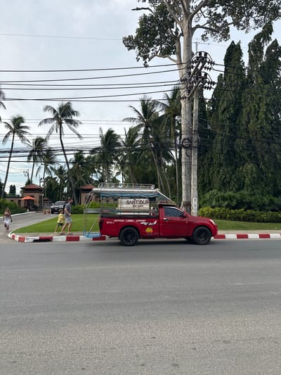 A red pickup truck with a covered seating area is parked on the right side of a paved road, facing right. The truck has a white and blue stripe on its side and "NATHON - LAMAI - AIRPORT" written on it. A man and a boy are walking on the sidewalk in front of the truck, heading away from the viewer. To the left of the truck, a woman in a white dress is walking towards the viewer. In the background, there are palm trees and buildings with orange roofs. The sky is overcast.