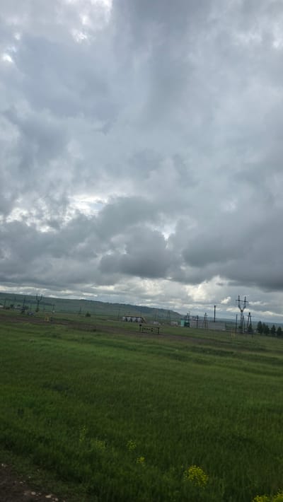 A wide-angle landscape photograph depicts a rural-industrial scene in Кама-Исмагилово, Russia, under a heavily overcast sky that fills the upper two-thirds of the frame with dense grey clouds. The mid-ground features a series of scattered industrial and utility structures, including multiple dark lattice-work and pole-type power pylons with visible overhead lines. Towards the center-right, a cluster of beige cylindrical tanks and associated piping, likely part of an oil or gas facility, is prominent. Various small, indistinct buildings, some light blue or grey, are interspersed among the infrastructure. A dirt track runs horizontally across the mid-ground. In the background on the left, low, undulating hills covered in green vegetation are visible. The foreground, occupying the bottom third of the image, is an expansive field of vibrant green, long grass, with scattered small patches of yellow flowers near the bottom. The overall lighting is soft and diffused, consistent with the overcast conditions, and no direct shadows are present. No people, animals, or explicit actions are depicted.