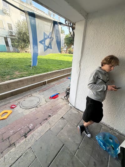 This image captures an outdoor residential scene in Migdal HaEmeq, Israel, under clear daylight conditions. A young male, appearing to be in his late teens or early twenties, with light brown, wavy hair, is positioned in the right foreground. He wears a grey hooded sweatshirt, black shorts, and black high-top sneakers, leaning against a textured, light-colored wall while focused on a black smartphone held in both hands. To his immediate right, a blue plastic bag containing multiple water bottles rests on the grey tiled ground. Extending to the left and into the midground is a paved walkway composed of reddish and greyish bricks, which includes a circular metal manhole cover. Several brightly colored plastic children's toys (yellow, red, and a blue/red object) are scattered across this walkway. A low concrete wall, topped by a white pipe, delineates the walkway from a vibrant green grassy lawn. In the upper left-to-midground, a large, slightly worn Israeli flag, white with two blue horizontal stripes and a blue Star of David, is suspended horizontally from an overhead line. In the background, light-colored residential buildings with visible air conditioning units and multiple satellite dishes are present, interspersed with green trees and foliage, all under a clear blue sky.