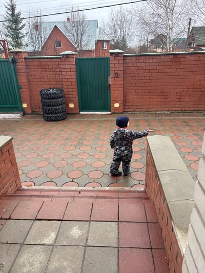 A child in a patterned jumpsuit and a dark blue hat stands on a tiled walkway. The walkway is made of patterned tiles with alternating grey and red circles. The child has their right arm raised. Behind the child, there is a brick wall with a green gate. Next to the gate, there are four stacked tires. In the background, there is a house and bare trees. The location is Shul'gino, Russia.