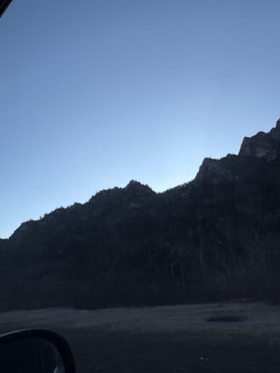 A stark, high-contrast image shows a jagged, dark cliff face silhouetted against a bright, clear sky. The landscape is rugged and imposing, with sharp edges defining the cliff's profile. A portion of a vehicle mirror is visible in the lower right corner of the shot, suggesting the image was taken from inside a car. The photograph is taken at an angle to give the view a different perspective.