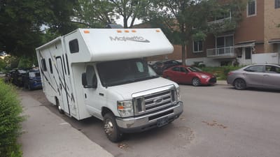 A white recreational vehicle (RV), identified by the "Majestic" logo on its upper section, is parked on the right side of a residential street in Montreal, Canada. The RV is the primary subject, occupying a significant portion of the foreground and midground. It is a Class C motorhome, characterized by the overhang above the driver's cabin. The RV is parked parallel to the curb, on a paved street with a concrete sidewalk.

Several other vehicles are also parked on the street. Immediately behind the RV, a dark-colored pickup truck and a blue SUV are visible. Further down the street, a red sedan and a silver sedan are parked. Beyond these vehicles, a brick building with multiple stories, featuring balconies and windows, is visible, indicating a residential neighborhood. Trees with lush green foliage line the street, casting shadows on the pavement. The weather appears to be overcast, with a hint of dusk in the sky. No people are clearly discernible in the image. The scene depicts a typical urban street in Montreal, with parked vehicles and residential buildings.