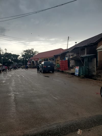 The image depicts a street scene in Barikin Gidar Kanat, Nigeria, under an overcast sky, suggesting either early morning or late afternoon. The road surface appears wet, indicating recent rain.
On the left side of the street are various shops and stalls, some with corrugated iron roofs. One stall has a prominent red sign that reads "PHARMACY." In front of the shops, stacks of red plastic crates are visible. A dark-colored SUV is parked by the roadside, and further down the street, a line of motorcycles suggests a mode of transportation or possibly a taxi rank. Several individuals are present, some walking along the street, others seemingly engaged in activities around the shops or near the parked vehicles. The overall impression is of a bustling, albeit somewhat cluttered, local market or commercial area.
The sky is a uniform gray, with no distinct sunlight. A few birds are visible in the sky, adding a touch of natural life to the urban setting. The shops appear to be constructed from a mix of wood and metal, with some exhibiting signs of age and wear. The presence of power lines running overhead indicates the availability of electricity in the area. The scene conveys a sense of everyday life and commerce in a Nigerian town.