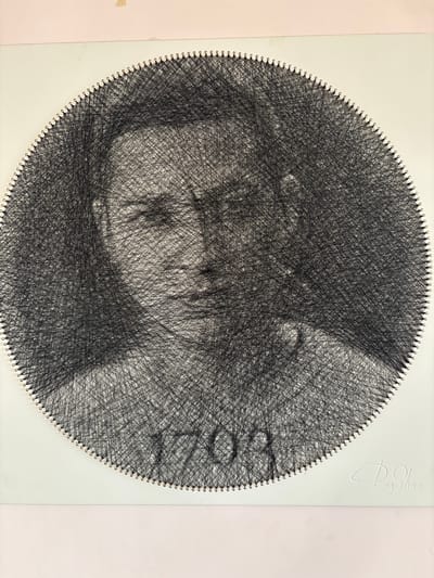 This is a string art portrait of a person, likely a woman, rendered within a circular frame. The artwork is created by stretching black thread between numerous small nails embedded in a white backing board. The varying density of the threads creates shading and defines the facial features, including eyes, nose, lips, and hair. The subject appears to be looking downwards, with a pensive or serene expression. The style is abstract and impressionistic, with the portrait emerging from the intricate network of threads.
In the upper right corner of the backing board, there is embossed text. The script is stylized and cursive, and appears to read "Ring Spring" or a similar phrase. The artwork is presented on a plain white surface, suggesting it is a photograph of the art piece itself, perhaps for display or sale. The lighting appears to be soft and diffused, with no harsh shadows, indicating an indoor setting. No specific location cues within Taraz, Kazakhstan are visible in the artwork or its immediate surroundings. The focus is entirely on the string art portrait.