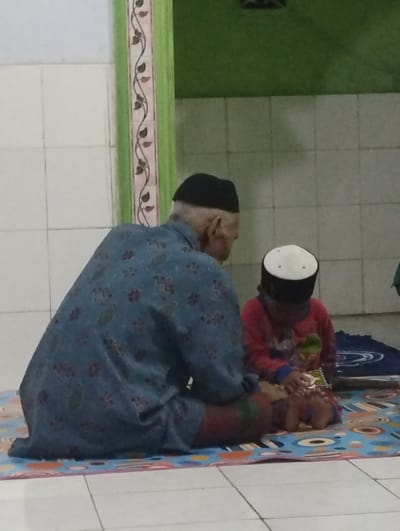 This indoor photograph captures an intimate scene between an older man and a young child, likely engaged in religious study or devotion, in what appears to be a mosque or a home in Kecamatan Arjasa, Indonesia. Both figures are seated on a colorful patterned mat laid over a tiled floor.
The older man, positioned on the left and viewed from behind, wears a black kopiah (Islamic prayer cap) and a loose, long-sleeved blue garment adorned with a repeating light-colored pattern. His lower body is covered by fabric that appears to be a dark red or maroon patterned sarong.
Beside him to the right, a young child, also seen from the back, is wearing a white and black kopiah and a red long-sleeved top. The child is holding a small book, suggesting they are reading or being instructed, possibly from a religious text such as the Quran. A folded dark blue prayer mat is visible on the floor next to the child.
The setting includes white square tiles on the lower part of the wall, topped by a section of plain green wall. A vertical decorative pillar or strip with a swirling vine pattern runs along the left side of the wall. The lighting is subdued, creating a quiet and focused ambiance for the activity. There is no visible text in the image.