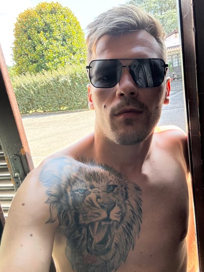 This is a portrait-style, close-up, eye-level, outdoor photo of a shirtless person with a large lion tattoo on their chest and shoulder. The person is wearing black sunglasses with a slight reflection of what appears to be an ornate interior in the lenses. They have short, light-colored hair and a light beard and stubble. Their skin is tanned, and there are a few small moles visible on their arm.
The background shows a bright, sunny outdoor scene with lush green foliage, likely trees or bushes, on the left side of the frame, and a lighter-colored paved or gravel area on the right. There is a dark wooden frame at the bottom and top of the image, suggesting the photo was taken through a window or doorway. The lighting indicates it is daytime, likely sunny.
The person appears relaxed, with their head tilted slightly back. The overall mood is casual and possibly candid. No visible text is present in the image. The location context is Sesto Calende, Italy.