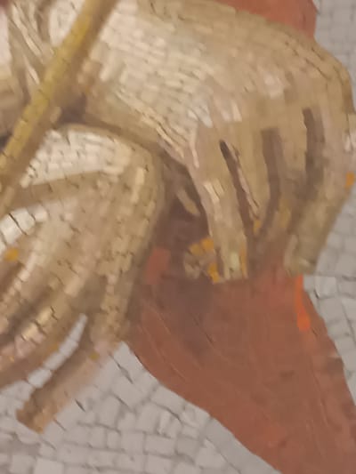 This is a close-up mosaic detail from the Ducal Palace of Colorno, Italy. The image focuses on two hands, rendered in a pale, creamy mosaic, holding a golden object, possibly a scepter or a scroll. The hands are detailed, showing distinct fingers and the texture of the skin. Behind the hands, a rust-colored mosaic forms a flowing shape, possibly representing drapery or a background element. The overall style suggests a classical or Renaissance depiction, common in Italian palaces. The mosaic tiles are small and tessellated, creating a somewhat pixelated appearance.