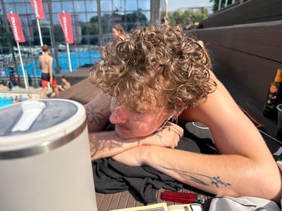 The image shows a young person with curly blonde hair lying down on a wooden deck. The person is wearing earphones and has tattoos on their arms. In the background, there is a swimming pool area with people visible. Red flags with white text are displayed in the background. The scene appears to be outdoors, likely at a public swimming facility. The location is Palilula Urban Municipality, Serbia.