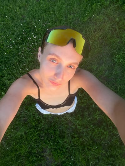 A high-angle, close-up, selfie-style shot shows a young person with blonde hair wearing reflective yellow sunglasses and a black bralette over white shorts, lying on a green grassy field. The person's face is tilted towards the camera, with their eyes open and looking upwards. The grass is lush and green, with small white and pink flowers interspersed throughout. The lighting suggests it is either early morning or late evening, casting a warm glow on the person's skin. The background is entirely covered by the grassy field. The image does not contain any information about the city of Подшивалово, Russia.