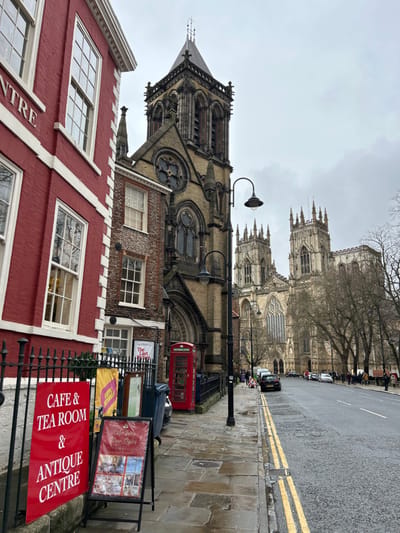 This image depicts a street scene in York, United Kingdom, specifically looking towards York Minster from Minster Yard or an adjacent street.
In the foreground to the left, a multi-story building constructed of red brick with white trim and numerous sash windows is partially visible, featuring a black metal fence. A large red sign attached to the fence advertises "CAFE & TEA ROOM & ANTIQUE CENTRE." An A-frame sign on the wet flagstone pavement displays "Cafe Tea Room Now Open."
Adjacent to the red brick building is a narrower, darker brick building with white window frames. Beyond this, St. Michael le Belfrey Church, a stone building with a tall spire and ornate Gothic-style architecture, including a prominent circular window, dominates the midground. A traditional red British telephone box stands on the pavement in front of the church's fence. A black street lamp with two lights is positioned centrally on the pavement.
In the background, the imposing Gothic structure of York Minster is clearly visible, featuring multiple large towers and intricate detailing. Bare deciduous trees line the street in front of the Minster.
The street, Minster Yard, is wet, indicating recent rain, and features double yellow lines. Several vehicles are parked along the street, and multiple pedestrians are visible on the pavements in the midground and background. The sky is uniformly overcast and grey.