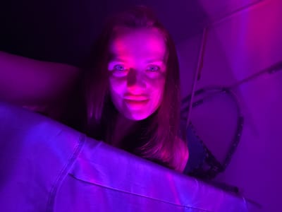 An overhead, medium shot of a young woman illuminated by purple and pink lights. She is looking directly into the camera with her mouth slightly open. Her hair is long and appears to be reddish-brown. She is leaning over a surface that is covered with a purple fabric. The background is dark with some objects visible, including what appears to be a rope or cord. The setting is indoors, likely a room, possibly in Rusalevka, Russia, though this cannot be definitively determined from the image alone.