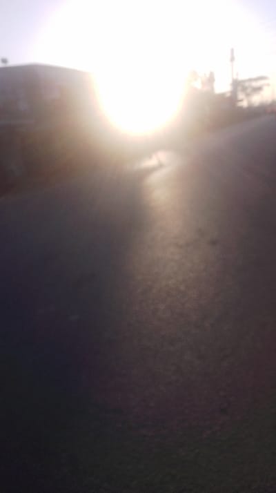 A blurry image shows a dark, textured road surface in the foreground, leading towards a bright sun or light source. The light overpowers the background details, creating a hazy effect. A building is seen to the left and some utility poles on the right, all rendered indistinct by the light. The scene evokes a sense of early morning or late afternoon sun on a road.