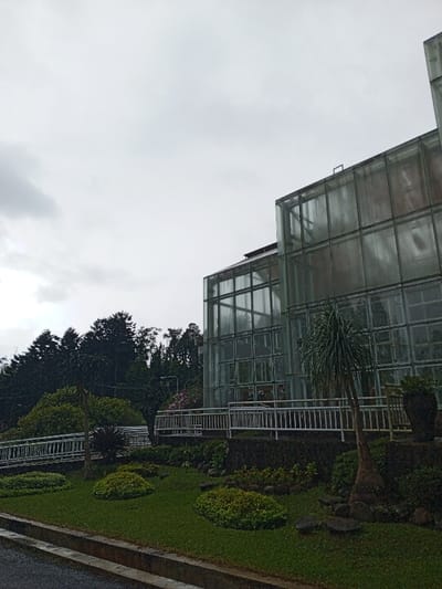 The image captures a modern glass-clad building set amidst lush natural surroundings under an overcast sky, likely in Khe Sanh Commune, Vietnam. The scene presents a tranquil outdoor environment with no apparent human activity.
The prominent subject is a large, multi-story structure with a facade composed of numerous square or rectangular translucent glass panels, giving it a greenhouse-like appearance. The glass reflects the gray sky and surrounding vegetation, indicating its reflective quality. Parts of the building's internal structure or dark roofing are visible through the glass at the top.
To the left of the building, a dense forest of dark green conifer-like trees rises up a hillside, suggesting a natural or mountainous landscape. In the foreground and around the building, manicured lawns are dotted with neatly trimmed bushes, smaller trees, and decorative rocks, creating a landscaped garden. A light-colored metal railing or fence curves along a pathway or elevated section of the garden, leading towards the building. A paved road or path with a curb is visible in the bottom left, suggesting accessibility.
The weather is distinctly overcast, with a uniform gray sky dominating the upper portion of the image, diffusing the light and creating soft shadows. This suggests either a cloudy day, early morning, or late afternoon. The overall mood is quiet and subdued. No people or visible text are present in the image.