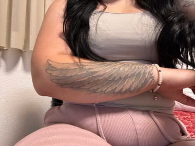 A close-up image captures a person, likely female, from the chest to the lap, in an indoor setting. The individual has long, dark hair that cascades over a light grey ribbed tank top. A prominent tattoo of a large wing, rendered in grey tones, covers the inner forearm. On the wrist, a white beaded bracelet featuring metallic accents and a small, bell-shaped charm is visible. The person wears light pink or mauve ribbed loungewear pants with an elasticated waistband. In the background to the left, light-colored vertical blinds or curtains are present, along with a textured white wall. To the right, a red patterned textile, possibly a rug or blanket, is visible. The lighting is soft and consistent with an indoor environment, suggesting a casual and relaxed atmosphere. The person's face is not shown, and no text is discernible in the image.