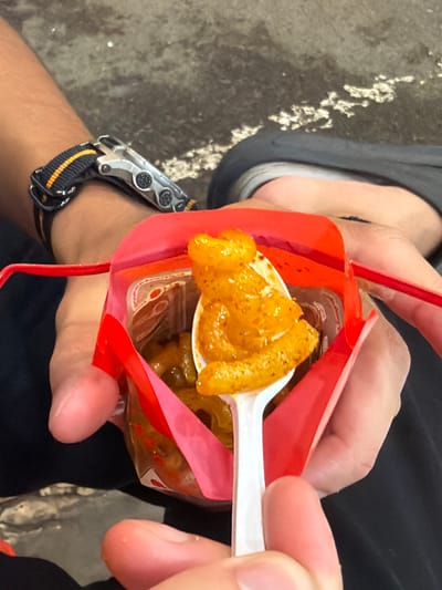 In a close-up shot, a person's hands are holding a red paper container filled with a spicy snack. A white plastic spoon is scooping up some of the snack, which appears to be small, crispy pieces coated in a vibrant orange sauce, possibly chili oil or a similar seasoning. The container itself has a clear plastic window, revealing more of the snack inside. One hand, with a watch on the wrist, holds the container open, while the other hand guides the spoon. The background is blurred, showing what appears to be a wet, textured surface, possibly pavement, and the dark fabric of clothing. The lighting suggests it's daytime. The setting might be outdoors, given the ground texture. The snack appears to be a popular street food item. The overall impression is casual, suggesting someone enjoying a snack on the go.