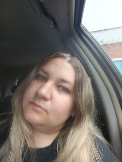 A woman is in the driver's seat of a car, taking a selfie. The car interior is dark, with a grey headliner. The woman has long, blonde hair and light blue eyes. She is wearing a black t-shirt. Through the open car window, a corrugated metal building and a brick building are visible, suggesting an industrial or commercial area in Meget, Russia. The sky is overcast.