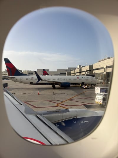 An aerial view through an airplane window shows several Delta Airlines airplanes parked at gates on an airport tarmac under a clear sky. The foreground features a portion of the wing of the airplane the photo is taken from. In the mid-ground, a Delta Airlines Boeing 737-900, registration N827DN, is parked at a gate. Another Delta Airlines plane is visible behind it. Various airport vehicles, including baggage carts and a tug, are present on the tarmac. In the background, airport terminal buildings are visible. The scene appears to be at Miami International Airport, as indicated by the presence of Delta Airlines, a major carrier at MIA, and the architectural style of the terminal buildings which is consistent with MIA.