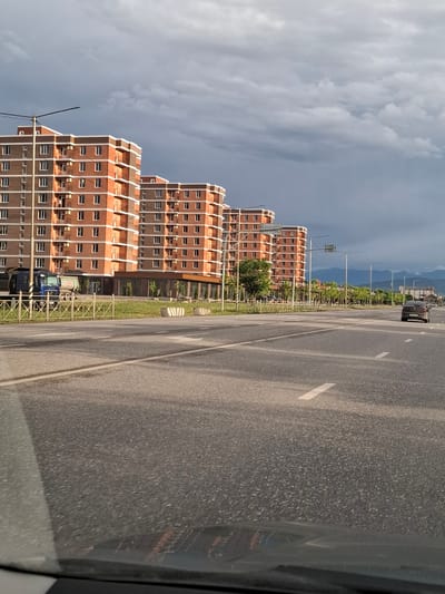 The image shows a street scene with modern, multi-story buildings made of red brick and gray concrete. The buildings are tall and have many windows. In the foreground, there is a paved road with white lane markings. A dark-colored van is parked on the side of the road. The scene appears to be in a developed urban or suburban area. The sky is overcast with gray clouds, suggesting a possibly late afternoon or early evening time. The presence of trees and a sidewalk area between the buildings and the road indicates a planned urban environment. The image is taken from a moving vehicle, as suggested by the slight blur and the perspective. The location is identified as Urus-Martan, Russia.